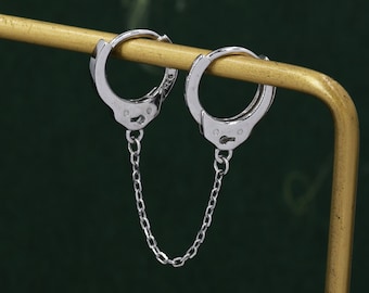 May include: Silver-colored handcuff earrings with a chain connecting the two hoops. The earrings are displayed on a gold-colored stand against a dark green background. The earrings have a polished finish and a detailed design.