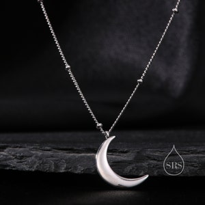 Puede incluir: Un collar de plata con un colgante de luna creciente y una cadena delicada con pequeñas cuentas. El collar se muestra sobre una superficie de piedra negra.