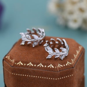 Op de afbeelding: Een paar zilveren oorringen in de vorm van een blad met heldere zirkonia-stenen. De oorringen worden gepresenteerd in een bruine fluwelen doos.