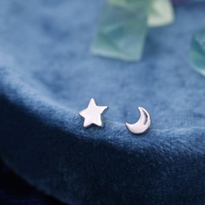 Puede incluir: Un par de pendientes de plata, uno con forma de estrella de cinco puntas y otro de media luna. Los pendientes se exhiben sobre una superficie de terciopelo azul, con unos pocos cristales transparentes, verde claro y azul en el fondo.