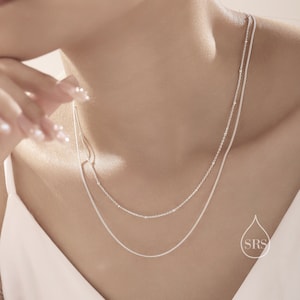 Peut inclure: Deux colliers en chaîne argentés superposés sur le cou d'une personne. La chaîne du haut a de petites perles, tandis que la chaîne du bas est une chaîne simple.