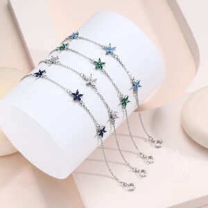 Può includere: Braccialetti in argento con ciondoli a forma di fiore in vari colori, tra cui blu, verde e bianco. I braccialetti sono esposti su una superficie bianca e curva, che ne mette in risalto il design delicato. Ogni braccialetto ha una catena con una chiusura.