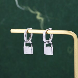 Puede incluir: Un par de pendientes de aro de plata con un dije de candado colgando de cada aro.