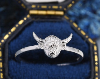 May include: A silver ring with a detailed Highland Cow head design. The ring is displayed in a blue velvet ring box.