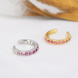 May include: Two ear cuffs with a row of pink and clear gemstones. The ear cuff on the left is silver and the ear cuff on the right is gold.