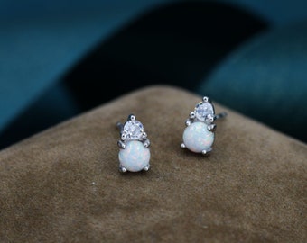 Puede incluir: Un par de pendientes de plata con una piedra de ópalo blanco y una piedra transparente más pequeña en la parte superior. Los pendientes se muestran sobre una superficie de terciopelo marrón.