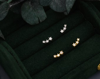 Puede incluir: Tres pares de pendientes con un diseño curvo adornado con pequeñas piedras de circonita cúbica transparentes. Los pendientes se muestran sobre un fondo de terciopelo verde.