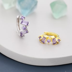 Könnte beinhalten: Zwei Creolen mit silberner und goldener Oberfläche. Beide Ohrringe haben ein Design aus lila Zirkonia-Steinen in verschiedenen Formen.