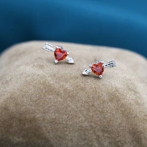 May include: A pair of silver earrings with red heart-shaped stones and arrow accents. The earrings are on a brown velvet surface.