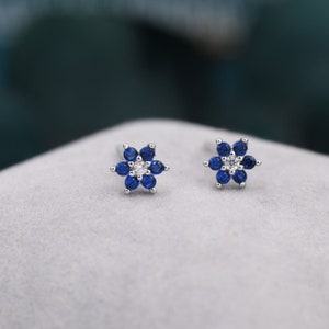 May include: A pair of flower-shaped stud earrings. Each earring features six deep blue gemstones surrounding a central white gemstone. The earrings are set in a silver-colored metal, creating a delicate floral design. The earrings are displayed on a light gray surface.