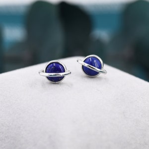 Puede incluir: Un par de pendientes de plata con una piedra preciosa azul en forma de planeta. El planeta está rodeado por un anillo de plata.