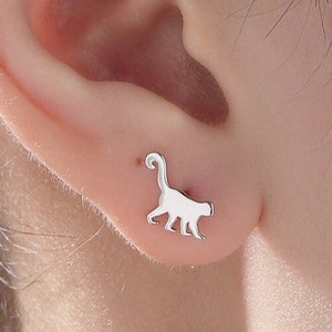 May include: A close-up of a person's ear with a silver monkey stud earring. The monkey is facing to the right and has a long tail.