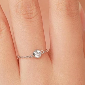 Op de afbeelding: Een delicate zilveren ring met een kleine, ronde, heldere edelsteen. De band van de ring is een fijne ketting, wat een minimalistische en elegante uitstraling geeft. De ring wordt getoond op een vinger, waarbij het ontwerp en de grootte worden benadrukt.
