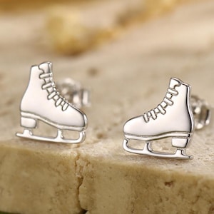 以下が含まれることがあります： 銀色のスケート靴スタッドピアス。ピアスは、レースとブレードを備えた、詳細なスケート靴の形をしています。ざらざらした明るい色の表面に表示されています。