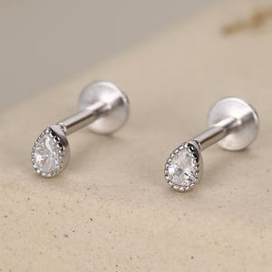 May include: Two silver-toned, teardrop-shaped earrings with a small, clear gemstone at the center of each. The gemstones are surrounded by a delicate, beaded border. The earrings have a flat, circular backing and a straight post.