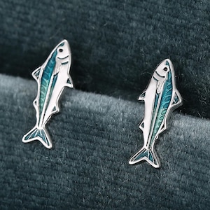 May include: A pair of silver-toned stud earrings shaped like small fish. Each earring features a detailed metallic outline with a vibrant teal and blue enamel inlay along the body, capturing the distinct form of a fish with fins and a tail.