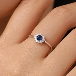 Può includere: Un anello in argento con una pietra preziosa centrale blu scuro a taglio rotondo, circondata da un alone di piccole pietre trasparenti a taglio rotondo. Il gambo dell'anello è sottile e lucido, con un delicato design ispirato a un fiore.