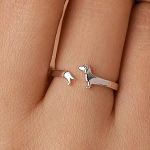 May include: A silver-colored ring featuring a dachshund dog design. The ring has an open design, with the dog's body and tail forming the ends of the band. The dog is depicted in a standing pose.