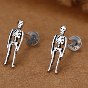 Può includere: Un paio di orecchini a lobo color argento a forma di scheletri umani. Ogni orecchino presenta una figura scheletrica dettagliata con teschio, cassa toracica e arti, fissata a un perno posteriore circolare piatto per un posizionamento sicuro.