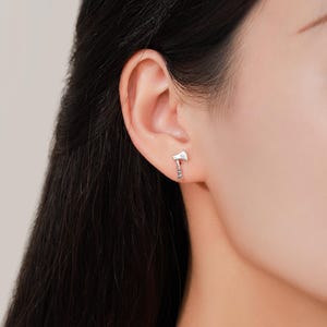 May include: A close-up of a silver axe earring. The earring is a small, detailed replica of an axe, with a blade and handle. The earring is worn on the ear of a person.