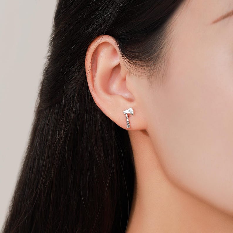 May include: A close-up of a silver axe earring. The earring is a small, detailed replica of an axe, with a blade and handle. The earring is worn on the ear of a person.