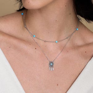 May include: A layered silver-tone necklace featuring two delicate chains. The upper chain is adorned with small, round, bright turquoise beads. The lower chain features a circular dreamcatcher pendant with a central turquoise bead and three small metal feathers.