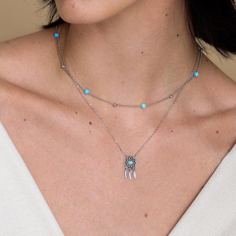 May include: A layered silver-tone necklace featuring two delicate chains. The upper chain is adorned with small, round, bright turquoise beads. The lower chain features a circular dreamcatcher pendant with a central turquoise bead and three small metal feathers.