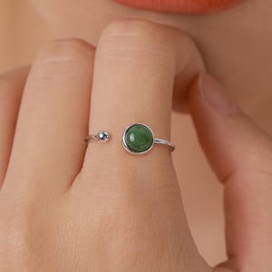 Op de afbeelding: Open zilveren ring met een ronde, levendige groene edelsteen en een kleine zilveren bal op de band. De ring is verstelbaar, met een opening tussen de edelsteen en de bal.