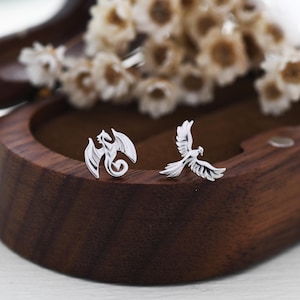 May include: A pair of silver-colored stud earrings. One earring is a dragon design, and the other is a bird design. The earrings are displayed on a dark wooden surface, with dried flowers in the background.