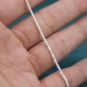 Puede incluir: Un collar de cadena de plata con un dise&ntilde;o texturizado y perlado. La cadena es fina y delicada, y se muestra en una mano.