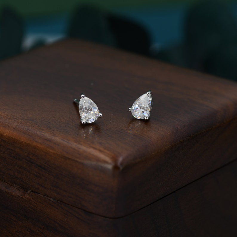 May include: A pair of stud earrings featuring pear-shaped, clear gemstones set in silver-colored metal. The earrings are displayed on a dark brown wooden surface, with a blurred background of teal and dark green.