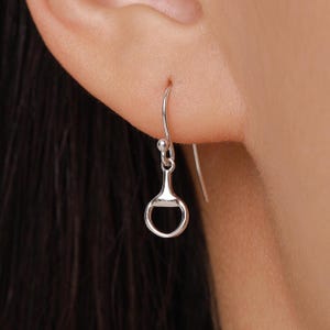Puede incluir: Un pendiente plateado con temática ecuestre. El pendiente presenta un pequeño y detallado diseño de bocado, suspendido de un alambre curvo para la oreja. El bocado tiene un anillo redondeado y una barra central. El pendiente se muestra en una oreja.
