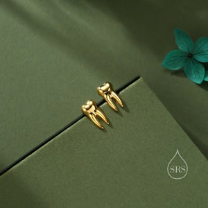 May include: A pair of polished gold-tone stud earrings shaped like human molars with distinct roots and a smooth, reflective surface. The earrings are positioned on a dark green surface next to a small logo featuring a water drop and the letters SRS.