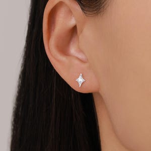 May include: A close-up of a silver starburst stud earring. The earring is small and has a detailed, textured design. The earring is worn on an ear, with the focus on the earring's shape and design.