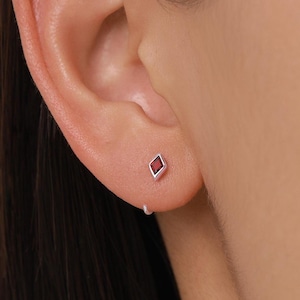 Puede incluir: Un solo pendiente de plata con una pequeña gema roja en forma de diamante. El pendiente tiene un diseño minimalista. El pendiente se muestra en una oreja.