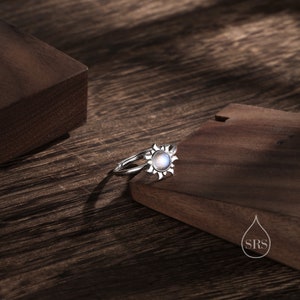 Puede incluir: Un anillo de plata con una piedra de luna engastada en un dise&ntilde;o de sol. El anillo est&aacute; sobre una superficie de madera.