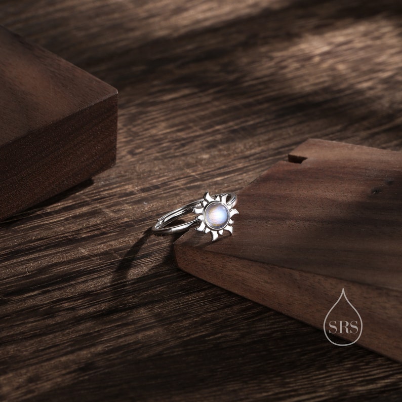 Puede incluir: Un anillo de plata con una piedra de luna engastada en un dise&ntilde;o de sol. El anillo est&aacute; sobre una superficie de madera.