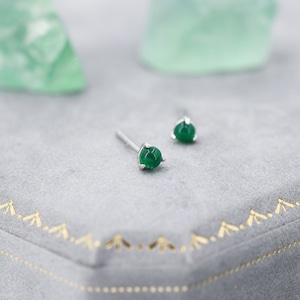 May include: A pair of stud earrings with round, vibrant green gemstones set in silver. The earrings are displayed on a gray surface with a gold decorative border, with a blurred green object in the background.