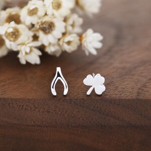 May include: A pair of silver stud earrings. One earring is shaped like a wishbone, and the other is a four-leaf clover. The earrings are displayed on a wooden surface with white flowers in the background.