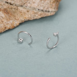 Peut inclure: Deux boucles d'oreilles ouvertes en argent, chacune avec une petite pierre pr&eacute;cieuse transparente de coupe carr&eacute;e &agrave; l'extr&eacute;mit&eacute; de la barre incurv&eacute;e. Les boucles d'oreilles sont pr&eacute;sent&eacute;es sur une surface bleu clair, avec une roche textur&eacute;e et beige en arri&egrave;re-plan.