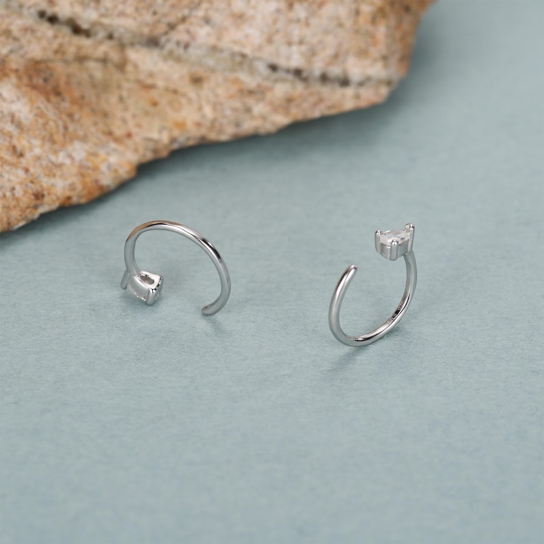Peut inclure: Deux boucles d'oreilles ouvertes en argent, chacune avec une petite pierre pr&eacute;cieuse transparente de coupe carr&eacute;e &agrave; l'extr&eacute;mit&eacute; de la barre incurv&eacute;e. Les boucles d'oreilles sont pr&eacute;sent&eacute;es sur une surface bleu clair, avec une roche textur&eacute;e et beige en arri&egrave;re-plan.
