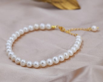 Peut inclure: Un collier de perles blanches avec un fermoir et une chaîne d'extension dorés. Le collier est sur un fond de tissu brun clair.