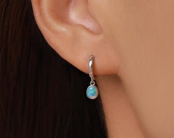 May include: A silver hoop earring featuring a small, oval-shaped blue opal charm. The earring is secured through the earlobe, with the polished metal hoop holding the iridescent blue stone pendant that displays flashes of green and light blue.