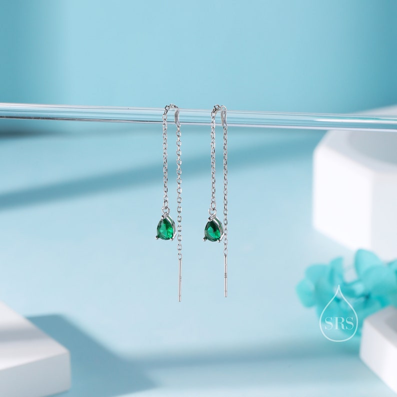 May include: A pair of silver threader earrings with teardrop-shaped, emerald-green gemstones. The earrings hang from delicate chains, creating a minimalist and elegant design. The background is a light blue.