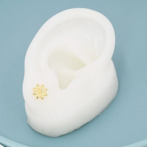 May include: A gold-colored star-shaped earring stud on a white ear model.