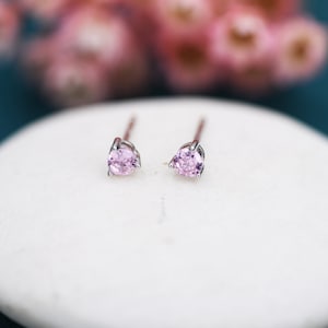 May include: A pair of stud earrings featuring light pink gemstones. The earrings have a silver setting and are presented on a white surface. The gemstones are round and pear-shaped. The background is blurred with pink and teal hues.