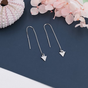 Può includere: Un paio di orecchini pendenti a triangolo d'argento con una catena delicata. Gli orecchini sono su uno sfondo blu.