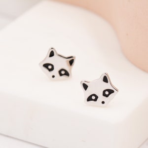 Pode incluir: Um par de brincos de press&atilde;o de prata com um design de guaxinim preto e branco.