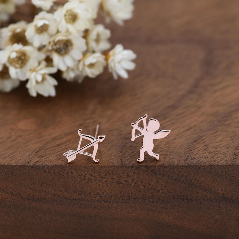 May include: A pair of rose gold stud earrings. One earring features an arrow and bow design, and the other depicts a small cupid figure with a bow and arrow. The earrings are set against a dark wood surface with white flowers in the background.