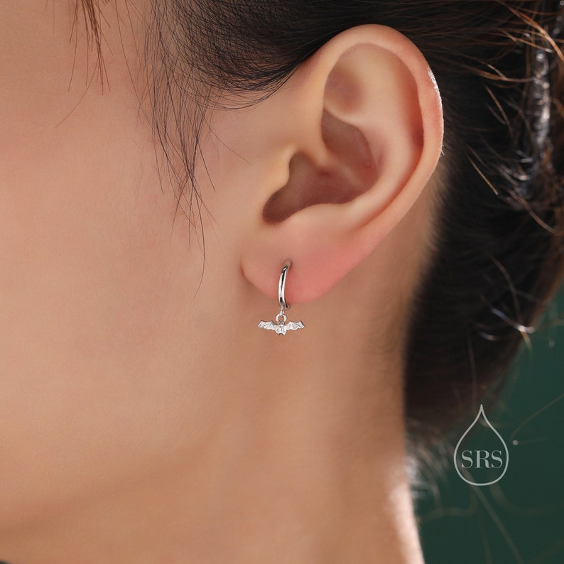May include: A silver hoop earring with a small bat charm hanging from it. The earring is worn in the left ear of a person.